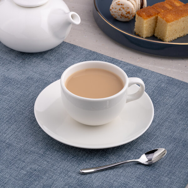 A warm white porcelain tea cup filled with tea, placed on a matching saucer with a spoon nearby.