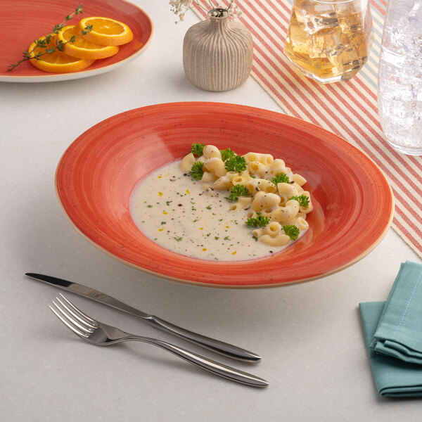 A red oval wide rim porcelain bowl filled with pasta and creamy sauce, garnished with herbs.