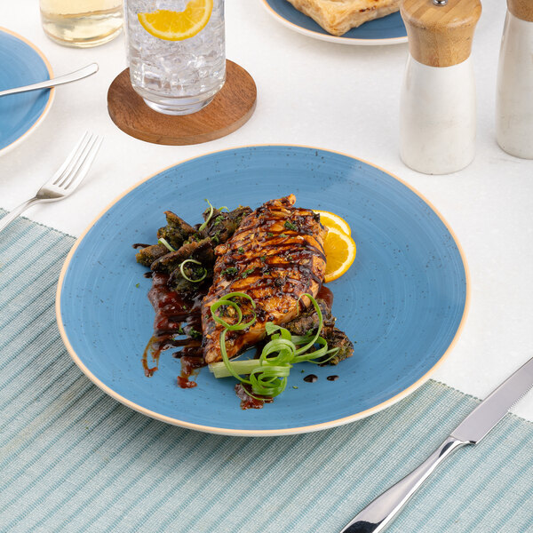 An oval blue porcelain steak plate with a grilled chicken breast, vegetables, and orange slices, set on a table with cutlery and a drink.