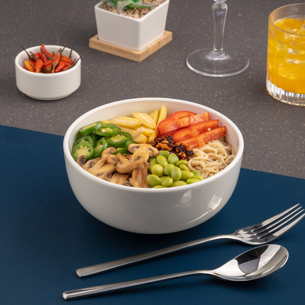 A white embossed porcelain bowl filled with assorted vegetables and noodles, placed on a table with cutlery.