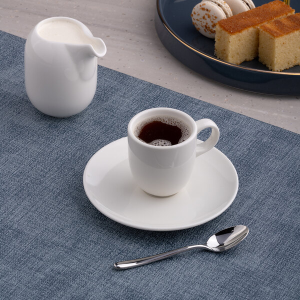 A small warm white porcelain espresso cup with a matching saucer, accompanied by a spoon and a milk jug, set on a blue placemat.