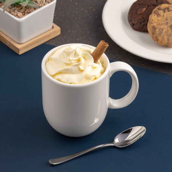 A white porcelain mug filled with a whipped topping and a cinnamon stick, placed next to a spoon.