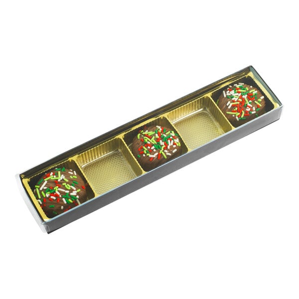 A rectangular brown and gold candy box with five cavities, containing three chocolate candies topped with red, green, and white sprinkles, and a transparent lid.