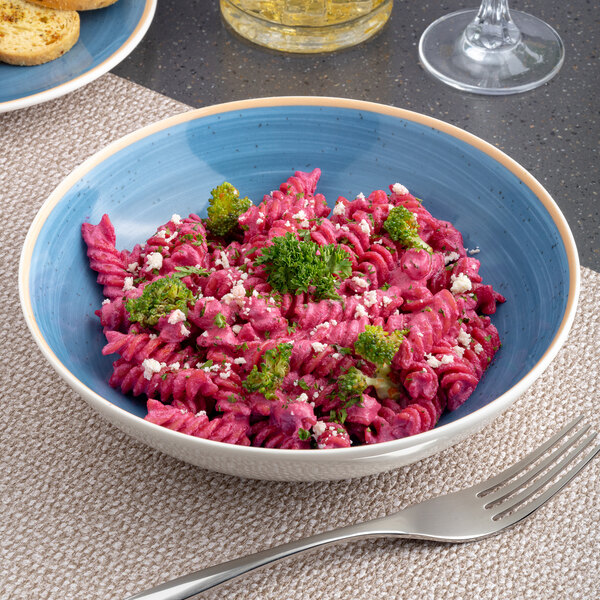 A blue and white two-tone round coupe deep porcelain plate filled with pink rotini pasta, broccoli, and garnished with herbs.