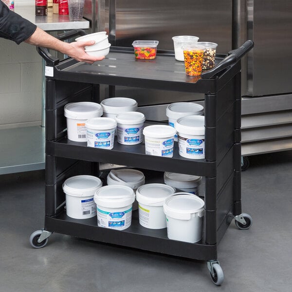 A person holding a Rubbermaid black bussing cart with containers of food on it.