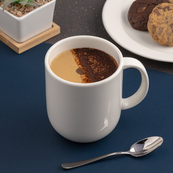 A warm white porcelain coupe mug filled with coffee, shown on a table with a spoon and cookies nearby.