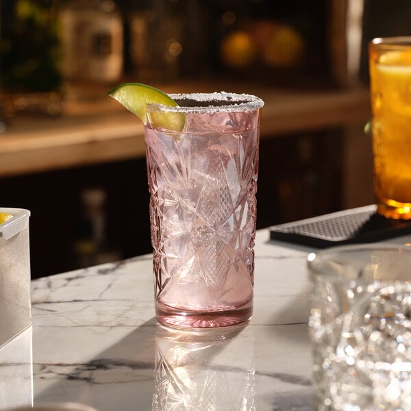 A pink patterned glass filled with ice and a drink, garnished with a lime wedge and a salted rim.
