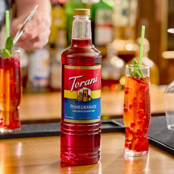 A 1-liter plastic bottle of Torani Pomegranate flavoring syrup displayed on a bar counter next to a glass of red beverage.