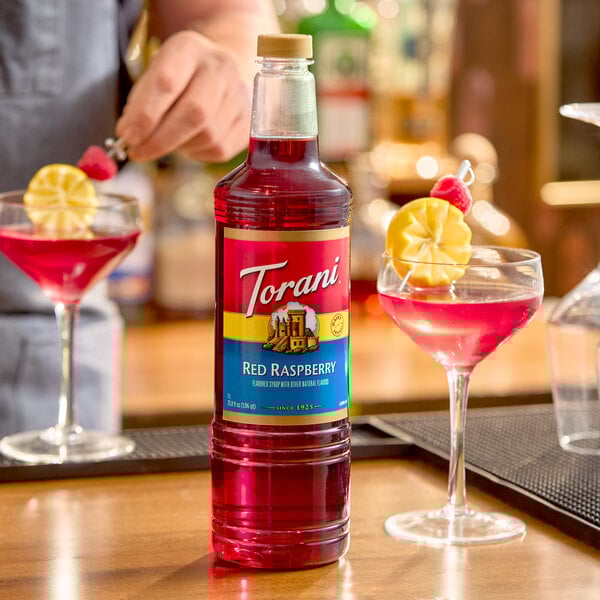 A 1-liter plastic bottle of Torani Red Raspberry flavoring syrup placed on a bar counter with two garnished cocktails nearby.