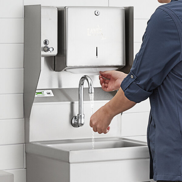 A person using a wall-mounted, hands-free stainless steel faucet and sink station with a paper towel dispenser.