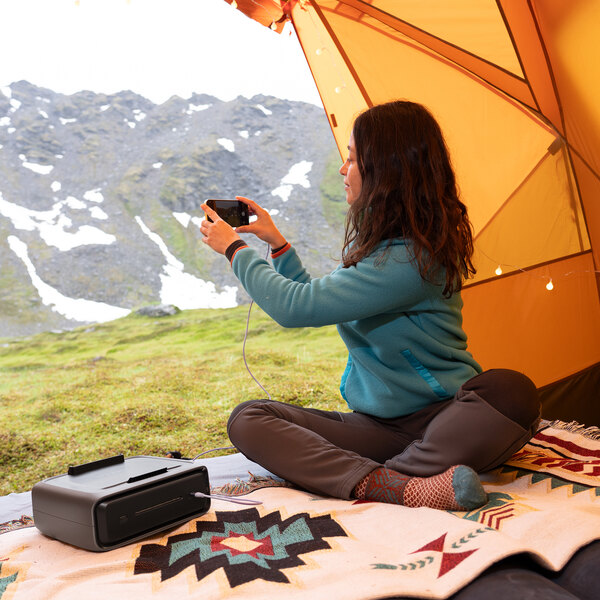 An EcoFlow RIVER 3 Plus EFEB300 lithium-ion extra battery placed on a patterned blanket inside a tent with a person using a device nearby.