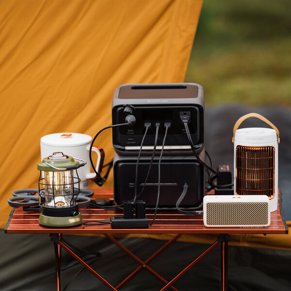A portable EcoFlow RIVER 3 Max Plus lithium-ion power station with an extra battery, shown powering various camping devices on a table.