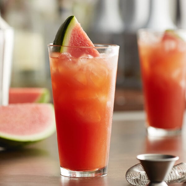 A tall, clear glass filled with a red beverage, ice, and garnished with a watermelon slice.