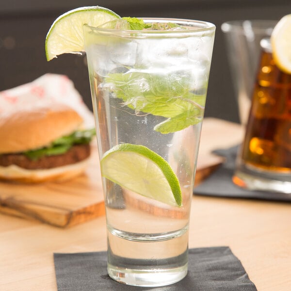 A clear highball glass filled with a beverage, ice, mint leaves, and lime wedges, placed on a black napkin.