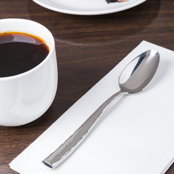 A stainless steel teaspoon with a textured handle placed on a white napkin next to a cup of coffee.