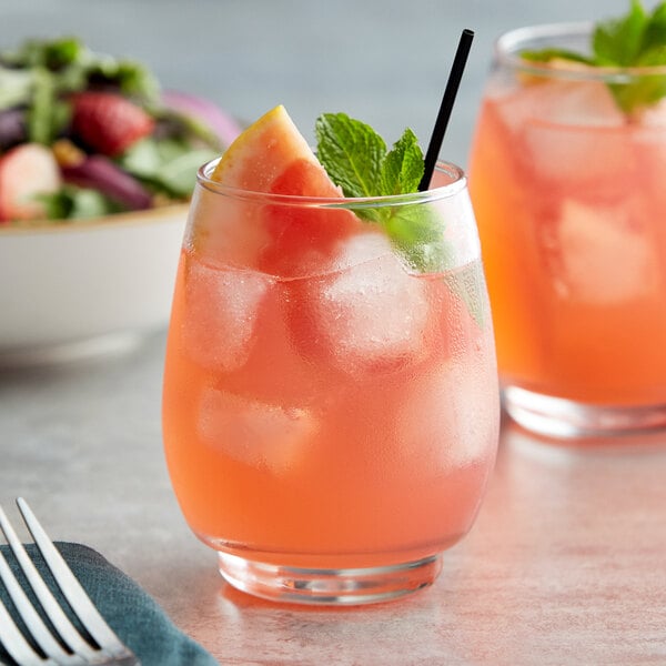 A clear, rounded 12 oz. stackable beverage glass filled with a pink drink, ice, a watermelon slice, and a mint garnish.