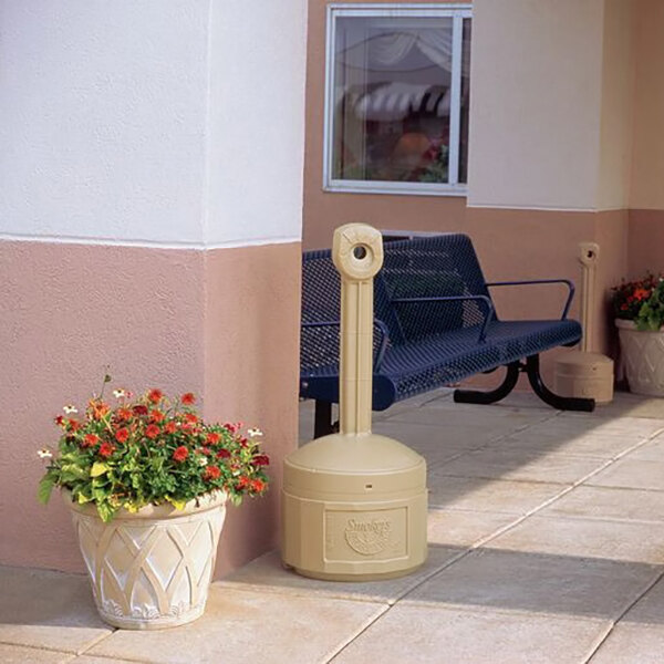 A beige plastic outdoor ashtray designed for smokers, placed on a sidewalk near a bench and a potted plant.