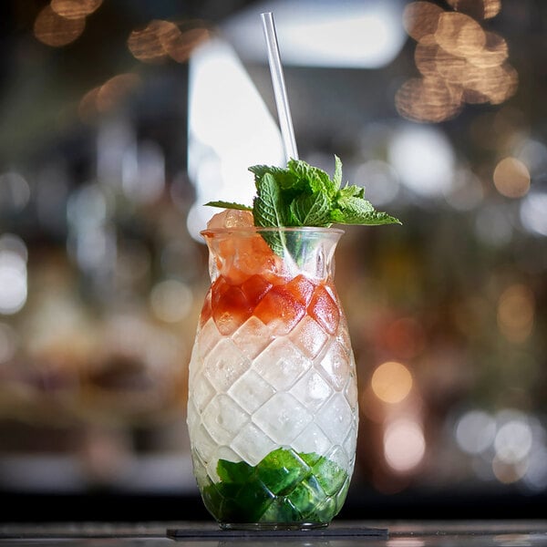 A clear, textured 17 oz. pineapple-shaped glass filled with a colorful cocktail, garnished with mint and a straw.