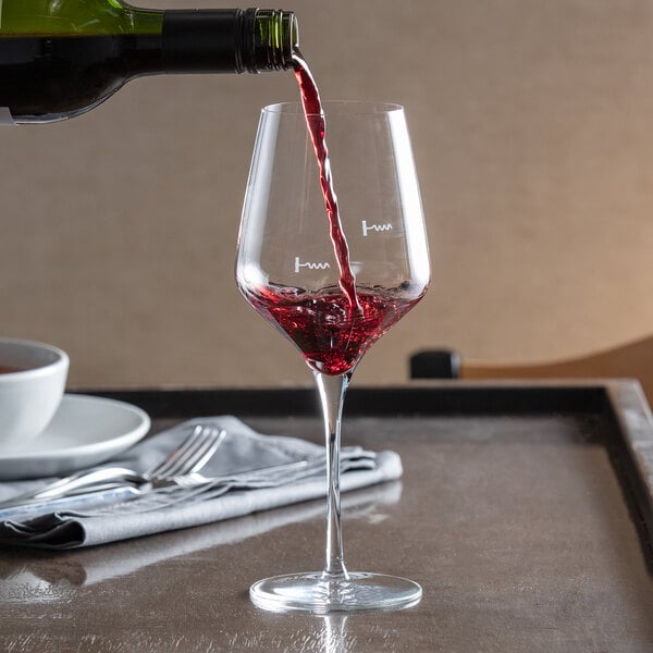 A clear wine glass with pour control markings being filled with red wine.