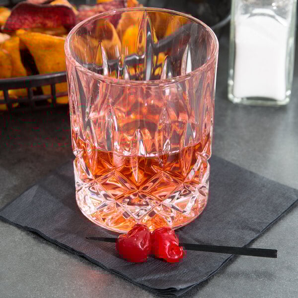A clear, faceted 10 oz. rocks glass with a reddish beverage, placed on a dark napkin with two maraschino cherries on a cocktail pick.
