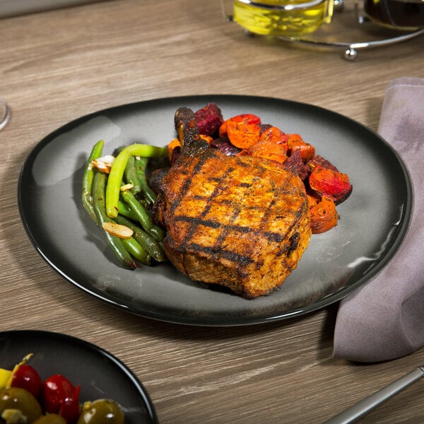 A dark gray porcelain plate with a grilled pork chop, green beans, and roasted vegetables.