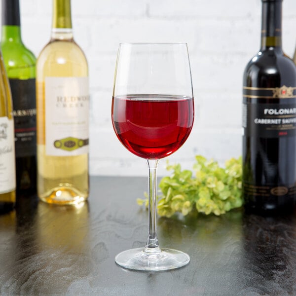 A clear 16 oz. wine glass filled with red wine, surrounded by bottles of wine on a dark surface.