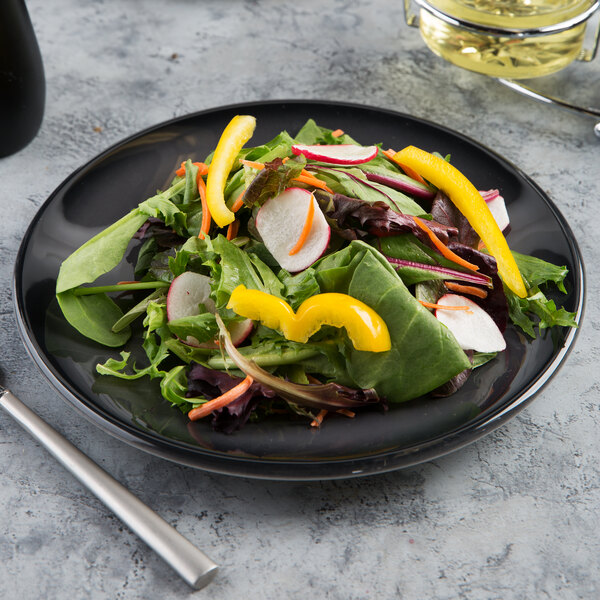 A black porcelain coupe plate with a fresh mixed green salad topped with sliced radishes, yellow bell peppers, and carrots.