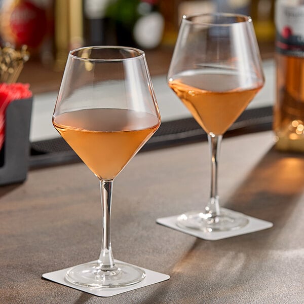 A pair of clear, stemmed wine glasses partially filled with rosÃ© wine, placed on square coasters on a bar surface.