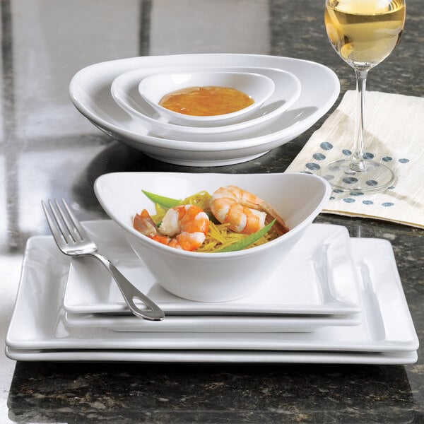 A small bright white oval porcelain bowl filled with food, displayed with matching plates and a glass of white wine.