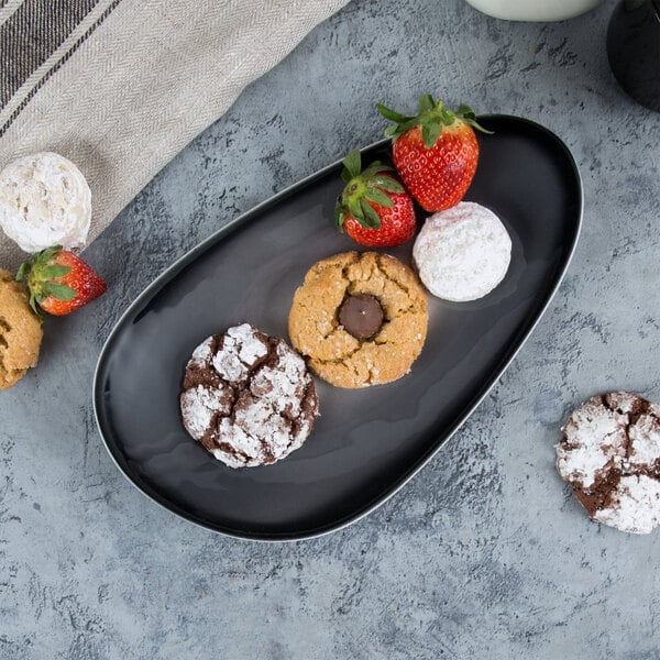 A black, oval-shaped porcelain tray displaying assorted cookies and strawberries.