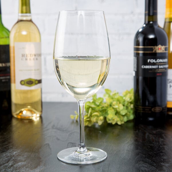 A clear 18 oz. wine glass filled with white wine, surrounded by bottles of wine on a dark surface.