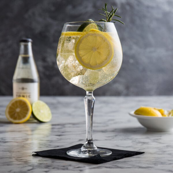 A large gin and tonic glass filled with a clear beverage, ice, lemon and lime slices, and a sprig of rosemary, placed on a black napkin on a marble surface.