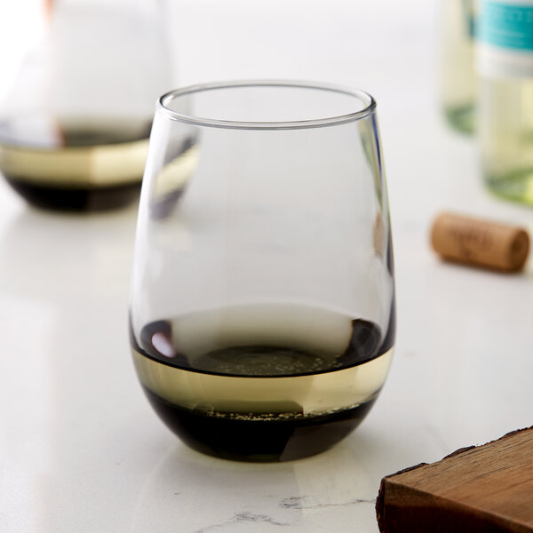 A clear stemless wine glass with a grey-tinted base, partially filled with white wine.