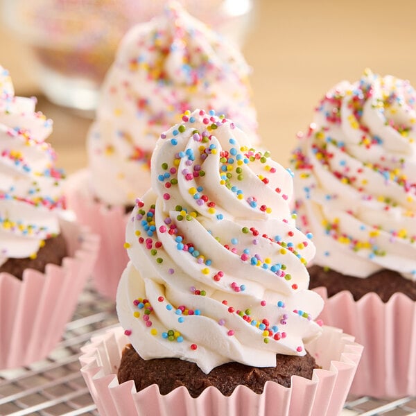 A chocolate cupcake with tall white frosting topped with colorful rainbow nonpareil sprinkles in a pink paper cup.