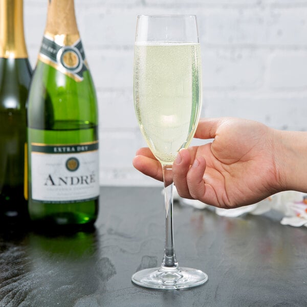 A clear 8 oz. champagne flute filled with sparkling wine, held by a hand, with bottles of champagne in the background.