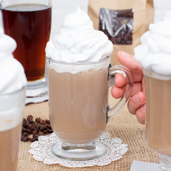 A clear glass coffee mug filled with a light brown beverage and topped with a generous amount of whipped cream.