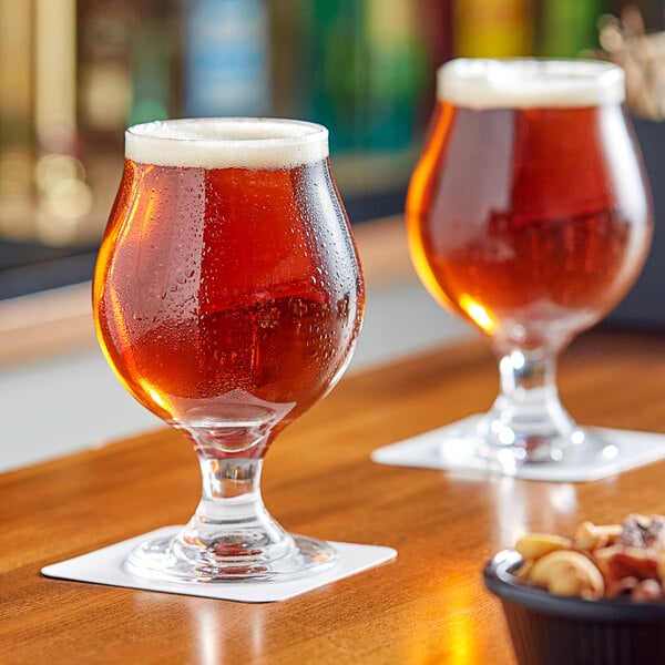 A 16 oz. Belgian beer tulip glass filled with amber beer, placed on a coaster on a wooden surface.