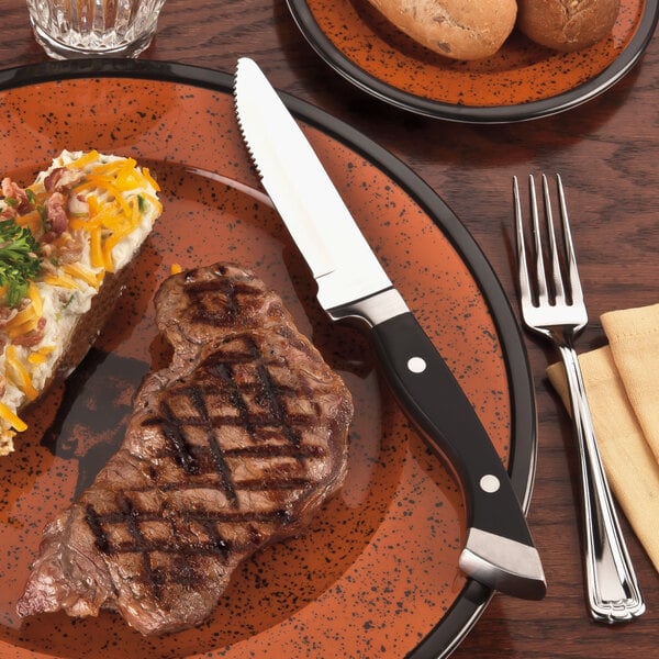 A stainless steel steak knife with a black polypropylene handle placed on a plate with food.