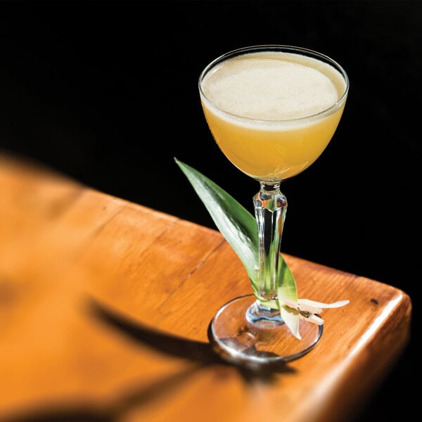 A clear Libbey Speakeasy 4.75 oz. Nick and Nora glass filled with a yellow cocktail and garnished with a green leaf and white flower.