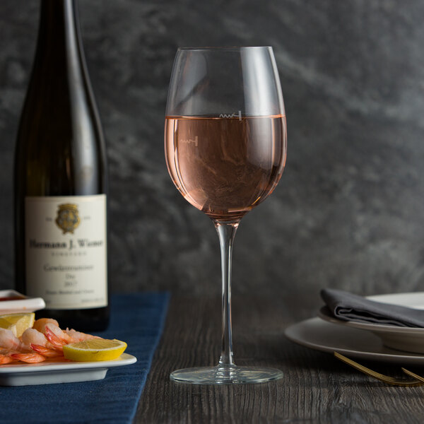 A clear wine glass with pour control markings, partially filled with rosÃ© wine, placed on a table set for dining.