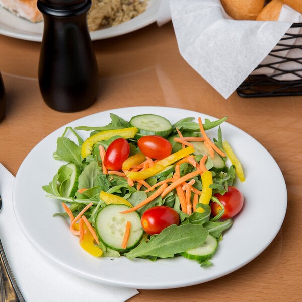 A white porcelain plate with a fresh salad containing arugula, cherry tomatoes, cucumber slices, shredded carrots, and yellow bell pepper strips.