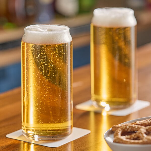 A clear 20 oz. can-shaped glass filled with light beer and topped with foam, placed on a coaster.