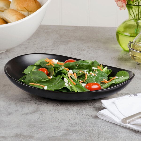 A black, oval-shaped porcelain coupe plate with a matte finish, shown holding a fresh salad.