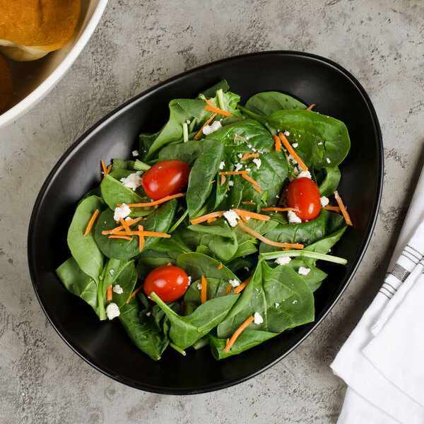 A black oval porcelain plate with a fresh spinach salad containing grape tomatoes, shredded carrots, and crumbled cheese.