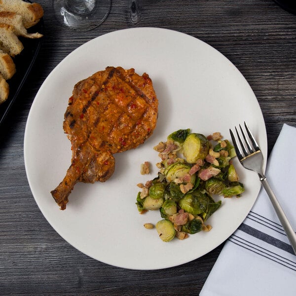 A large round white porcelain coupe plate with a grilled pork chop and a side of Brussels sprouts with bacon.