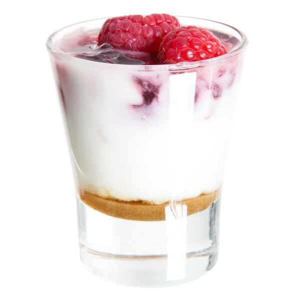 A small clear glass filled with layered dessert, including a crumb base, creamy filling, and raspberries on top.