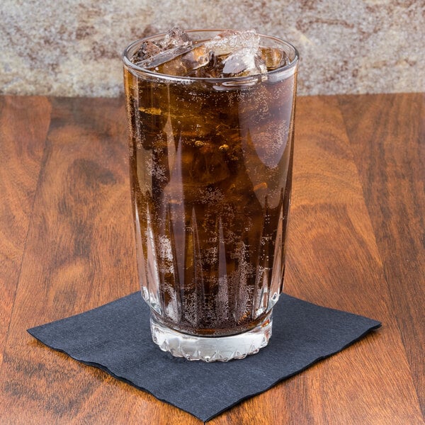 A clear 16 oz. cooler glass filled with a dark carbonated beverage and ice, placed on a black napkin on a wooden surface.