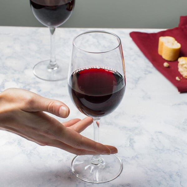 A clear 12 oz. wine glass filled with red wine, shown on a marble surface with a hand holding the stem.