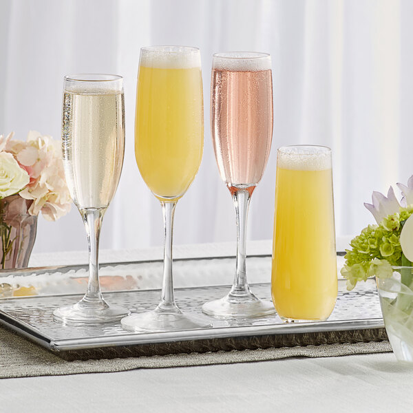 A clear 8.5 oz. stemless flute glass filled with a yellow beverage, displayed alongside three stemmed flute glasses on a tray.