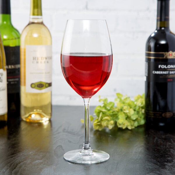 A clear 17 oz. wine glass filled with red wine, surrounded by bottles of wine on a dark surface.